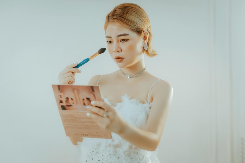 An Asian woman with short hair applies makeup using a brush while looking in a mirror.