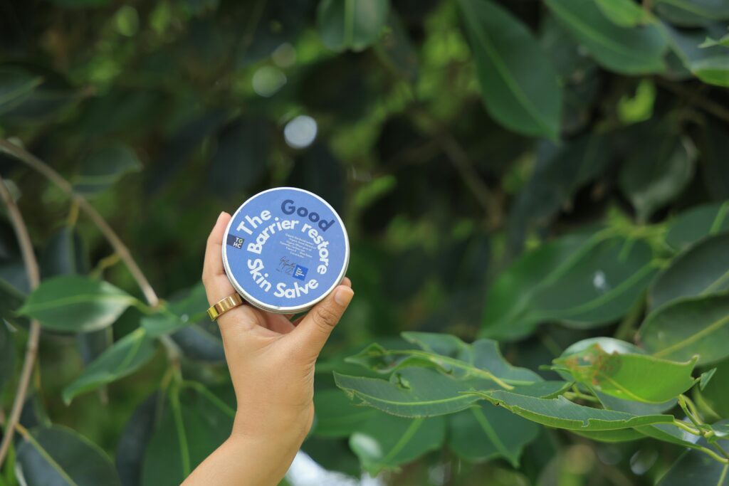 A hand holds a skincare salve against a lush green leaf background.