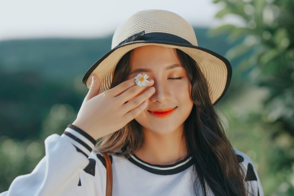 Close-up portrait of a smiling woman in a straw hat, holding a daisy over one eye against a blurred outdoor background.