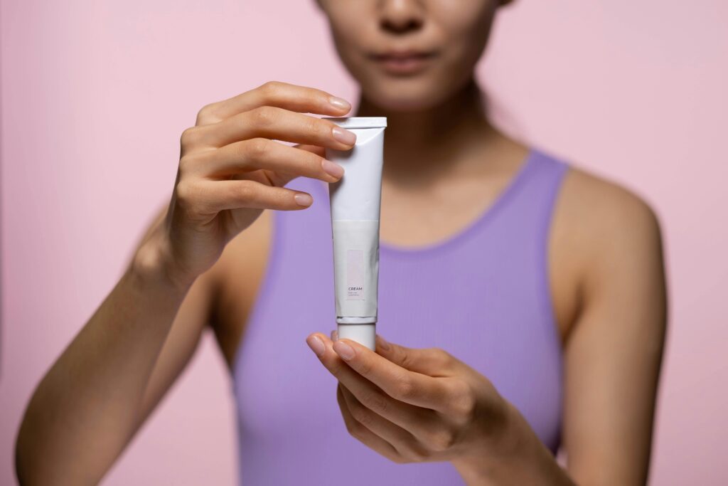 A woman in a purple tank top holds a skincare cream against a pink background in a studio setting.