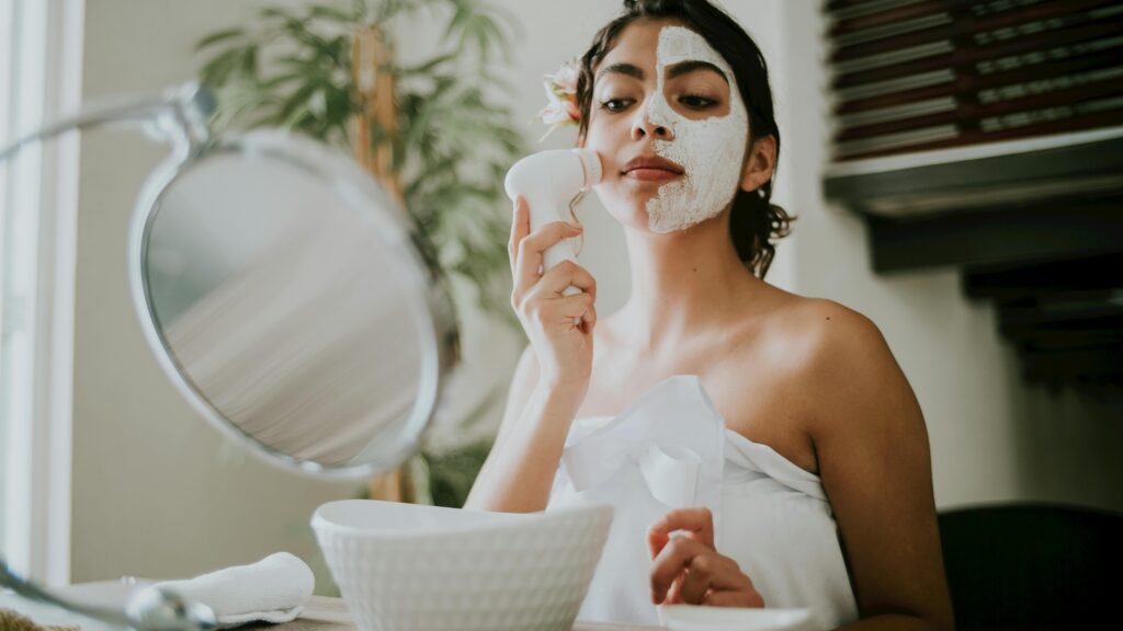 A woman uses a facial brush to apply a clay mask during a relaxing skincare routine.